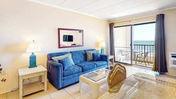 A room with a blue sofa, a glass coffee table, a wicker chair, a TV mounted on the wall, and a balcony with a view of the sea. - Eastwinds 505 (Ocean City)