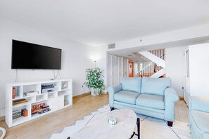 A living room with a flat-screen TV, a light blue sofa, a white entertainment unit, and a staircase leading to another level. - Oceanfront Century I Condo w/ Ocean View, WiFi, Washer/Dryer, AC, Shared Pool (Ocean City)