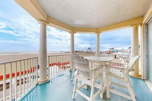 A covered outdoor area with a table and chairs, a railing, and a view of the beach and distant structures. - Belmont Towers 301 (Ocean City)