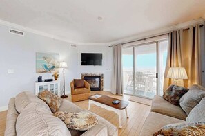 Ein Wohnzimmer mit einer Sofagarnitur, einem Couchtisch, einem Kamin, einem Fernseher und einem Balkon mit Vorhängen.