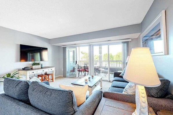 A modern living room with a sofa set, a coffee table, and a TV. There is a balcony with a view of greenery. - Heron Haven (Ocean City)