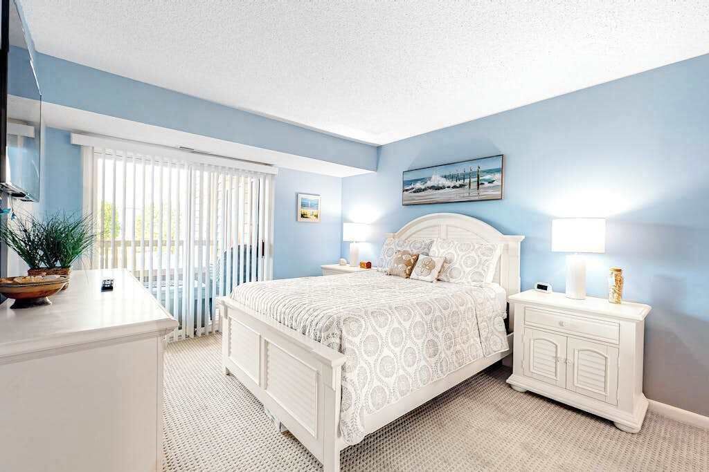 A bedroom with a bed, bedside tables, a nightstand, a lamp, a window with blinds, and a wall-mounted picture.