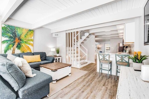 A modern living room with a sofa, coffee table, and a painting of a palm tree. - Spacious townhouse with ocean view, private beach area, pool access, W/D & AC (Ocean City)