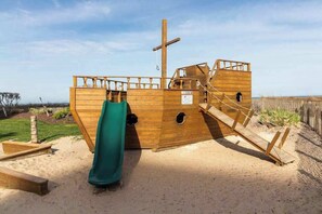 A wooden pirate ship play structure with a slide, stairs, and a flagpole. - Carousel 2105 (Ocean City)