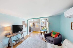 Ein Wohnzimmer mit Fernseher auf Holzständer, einer Couch, einem Esstisch und Blick zur Küche.
