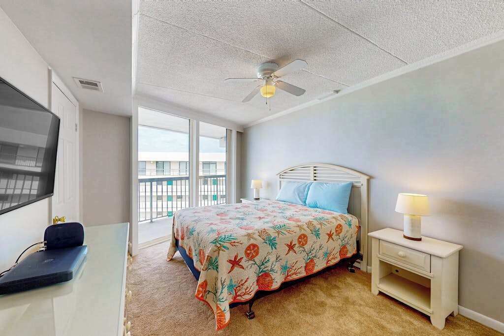 Un dormitorio con cama, ventilador de techo, televisor, mesita de noche y ventana con vistas al balcón.