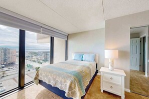 Een slaapkamer met een groot raam, een bed met een geborduurd dekbed, een nachtkastje met een lamp en een uitzicht op een stadsgezicht.