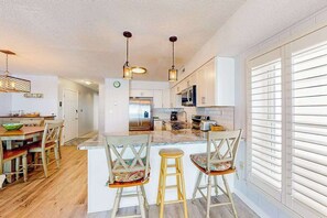 A kitchen with a central island, bar stools, a dining area with a wooden table and chairs, and a view of the outside through large windows. - Ultimate beach getaway with ocean view, open layout, garage parking, & streaming (Ocean City)