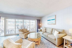 A living room with a sofa, armchairs, a glass coffee table, and a view through large windows to an outdoor balcony. - Ultimate beach getaway with ocean view, open layout, garage parking, & streaming (Ocean City)