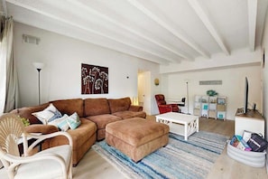 Ein Wohnzimmer mit braunem Sofa, weißem Sessel, Hocker und einem weißen Couchtisch. Ein blau-weiß gestreifter Teppich, eine weiße Decke und ein an der Wand befestigtes Kunstwerk sind ebenfalls vorhanden.