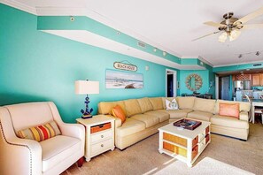 Un salon avec un mur de couleur turquoise, un canapé d’angle beige, un fauteuil blanc, une table basse et un ventilateur de plafond.