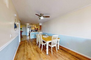A dining area with a wooden table and chairs, a ceiling fan, and a kitchen in the background. - San Remo 102 (Ocean City)