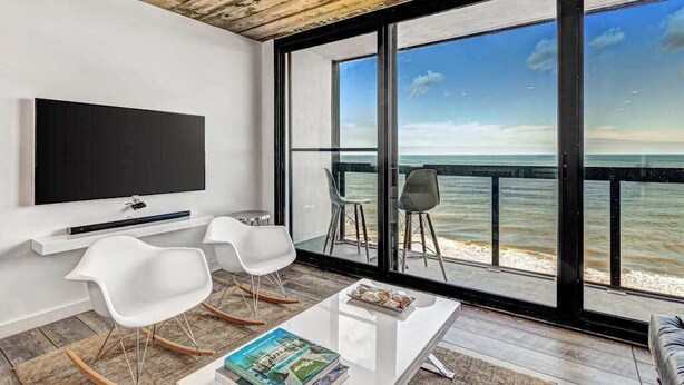 Ein modernes Zimmer mit Fernseher, weißen Stühlen, einem Couchtisch und einem großen Fenster mit Meerblick.