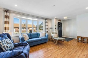 A spacious living room with a blue sofa, a wicker chair, and a large window offering a view of the outside. - Spacious Family Home with Full Kitchen & Deck - Close to the Beach (Ocean City)