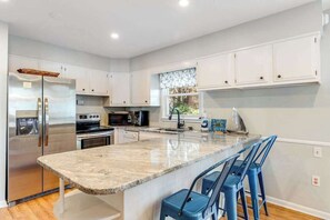 A modern kitchen with white cabinets, stainless steel appliances, a marble countertop, and blue bar stools. - Spacious Family Home with Full Kitchen & Deck - Close to the Beach (Ocean City)
