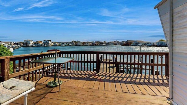 Una terraza de madera con una mesa y sillas, con vistas a un cuerpo de agua y edificios al fondo.