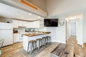 A modern kitchen with white cabinets, a wooden island with bar stools, a flat-screen TV mounted on the wall, and a wooden floor. - La Vista Bay 112 and 114 on 75th St Combo -A,B (Ocean City)
