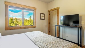 Quarto com cama, televisão sobre suporte, janela com vista para árvores e colinas, e porta.