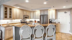 A kitchen with white cabinetry, a granite countertop, a stainless steel refrigerator, and three white chairs with pineapple designs. - Belmont Towers 608 (Ocean City)