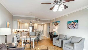 A modern living room with a glass coffee table, grey sofas, a dining area with a white table and chairs, and a kitchen in the background. - Belmont Towers 608 (Ocean City)