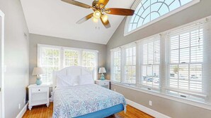 Un dormitorio con cama, dos mesitas de noche, ventilador de techo y ventanas amplias.
