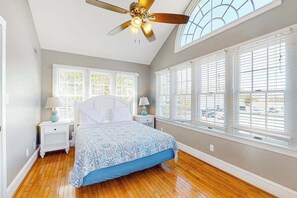 Ein Schlafzimmer mit einem Bett, zwei Nachttischen, einem Deckenventilator und großen Fenstern.
