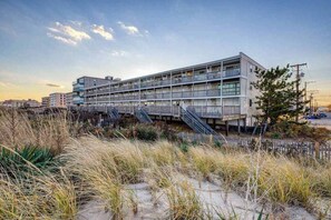 A multi-story building with balconies, a staircase, and a sandy foreground with grass. - Oceanfront condo with beach access, spacious deck, full kitchen & private W/D (Ocean City)