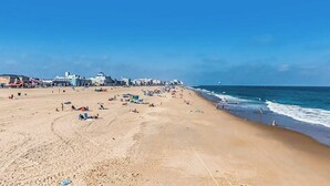 Ein weiter Sandstrand mit Sonnenschirmen und Badegästen, ein klarer, blauer Himmel und Gebäude im Hintergrund.