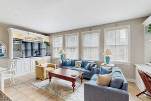Ein Wohnzimmer mit Sofa, Couchtisch und Fernseher. Es gibt Fenster mit Jalousien und eine Pflanze auf der Fensterbank.