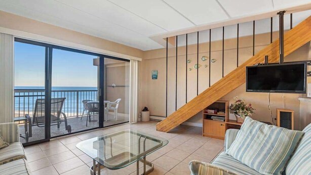 A spacious living area with a glass coffee table, a sofa, a TV mounted on the wall, and a balcony with a view of the sea. - High Point South P10 (Ocean City)