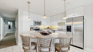A modern kitchen with white cabinets, a stainless steel refrigerator, and a central island with bar stools. - Gateway Grand 507 (Ocean City)