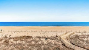 Een strand met een houten omheining, zandduinen en een helderblauwe lucht.