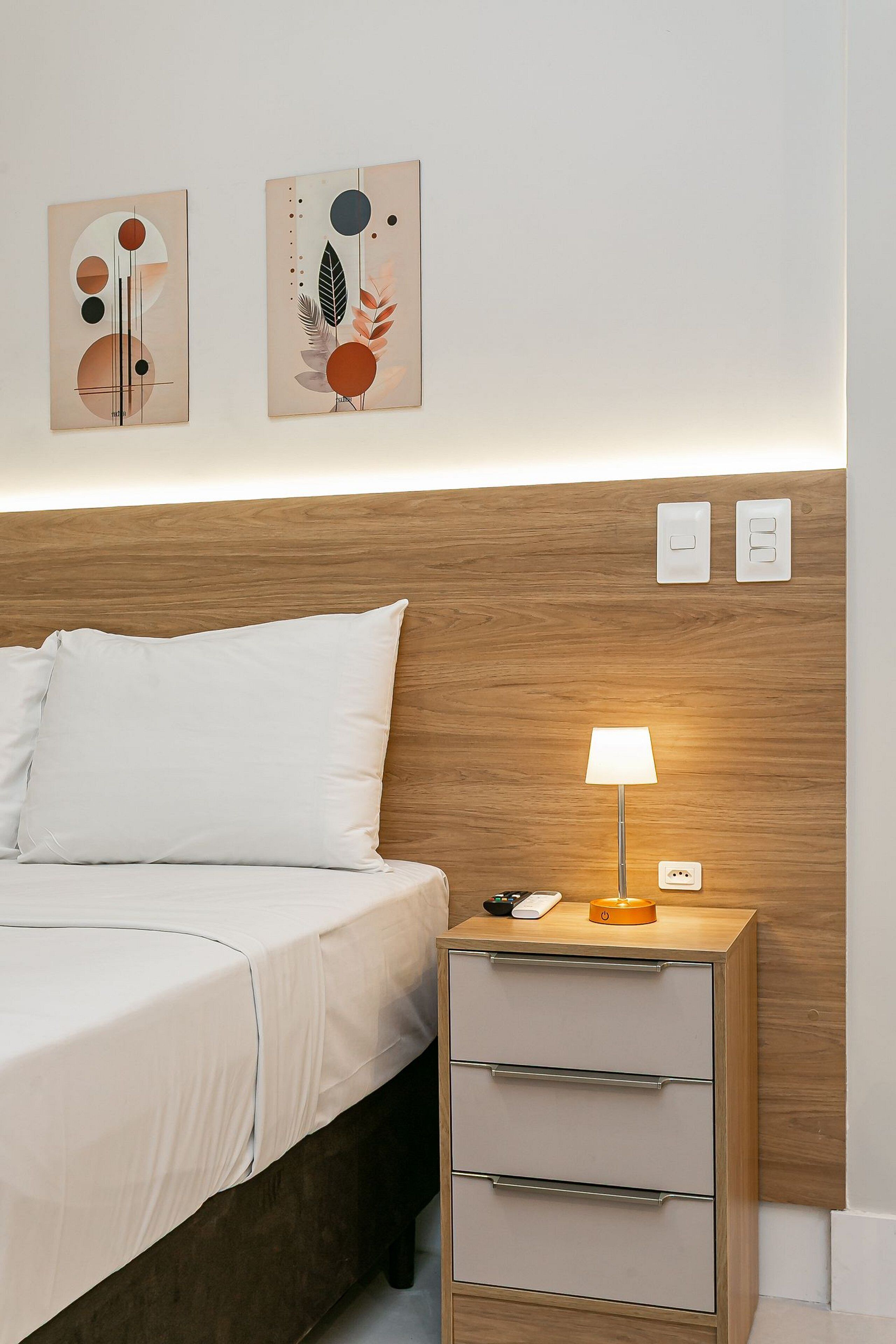 Un letto rifatto con lenzuola bianche, una testiera in legno, un comodino con una lampada e due interruttori a parete.