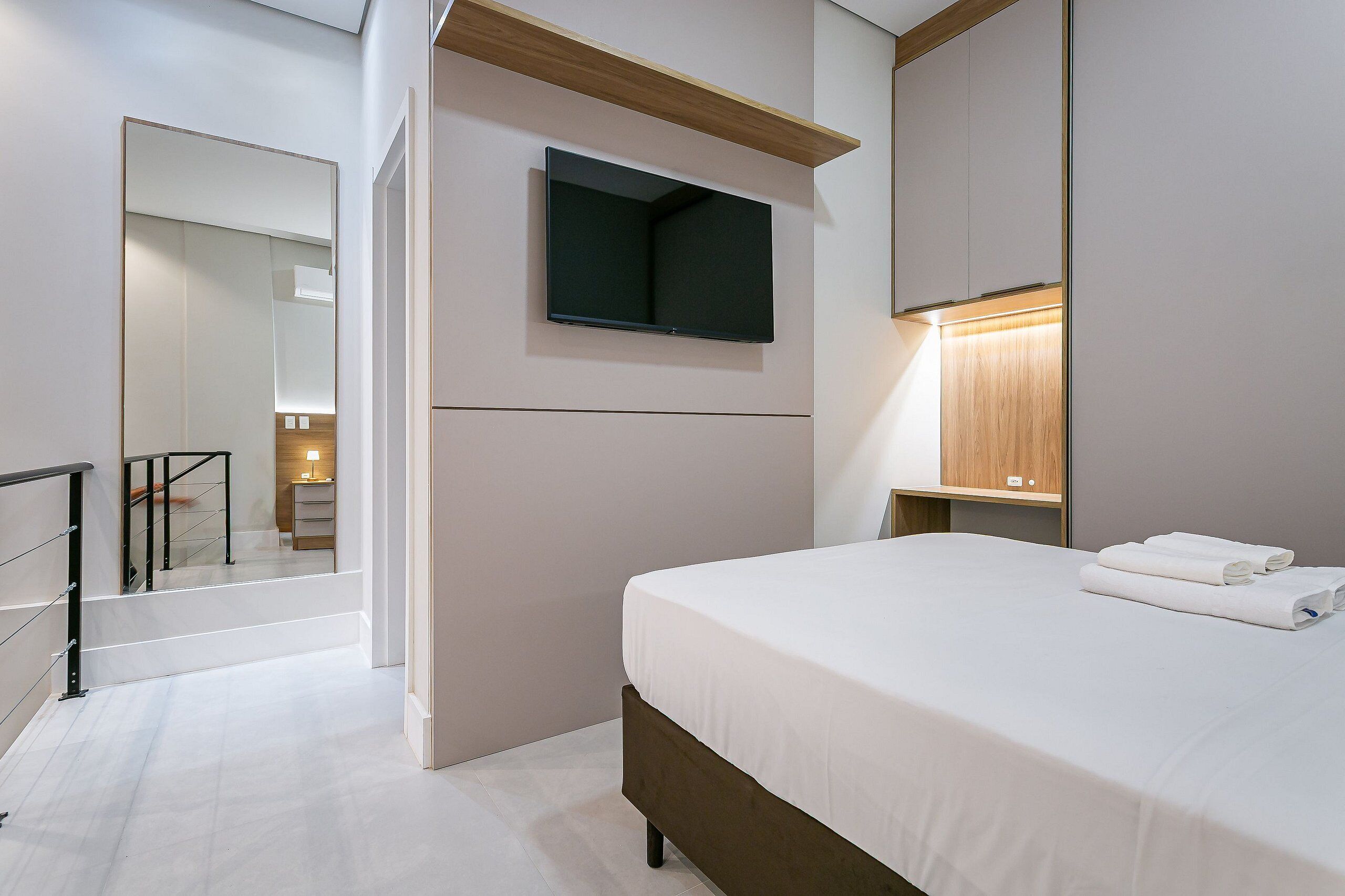 A modern hotel room with a bed, a flat-screen TV mounted on the wall, a bedside table with a lamp, and a view of the hallway.
