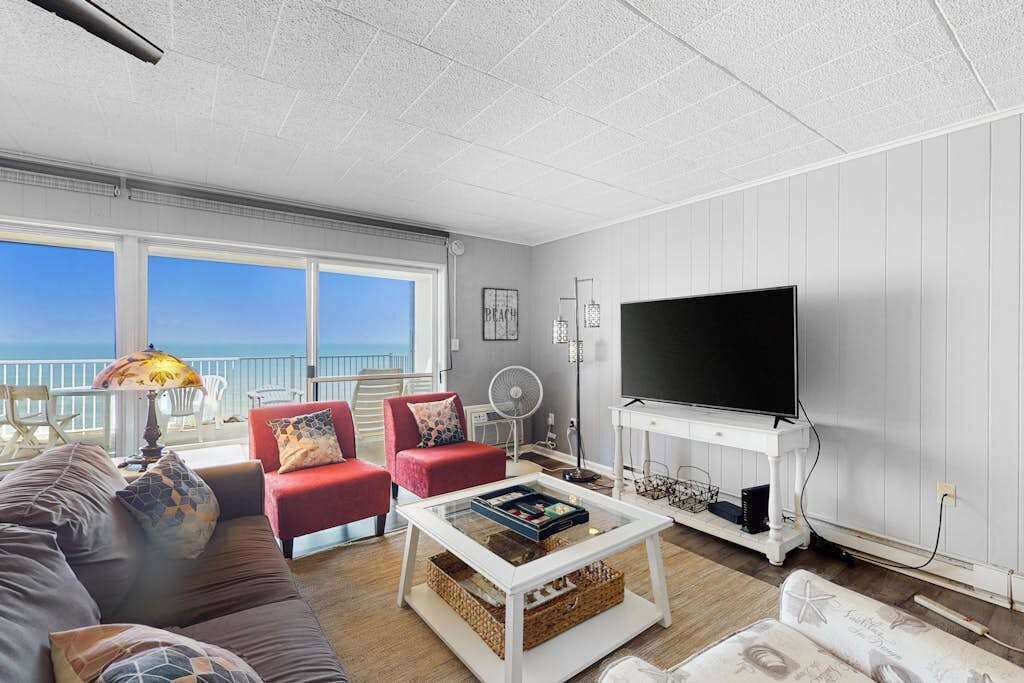 Ein Wohnzimmer mit einer Couch, roten Sesseln, einem Couchtisch, einem Fernseher und einem Balkon mit Blick.
