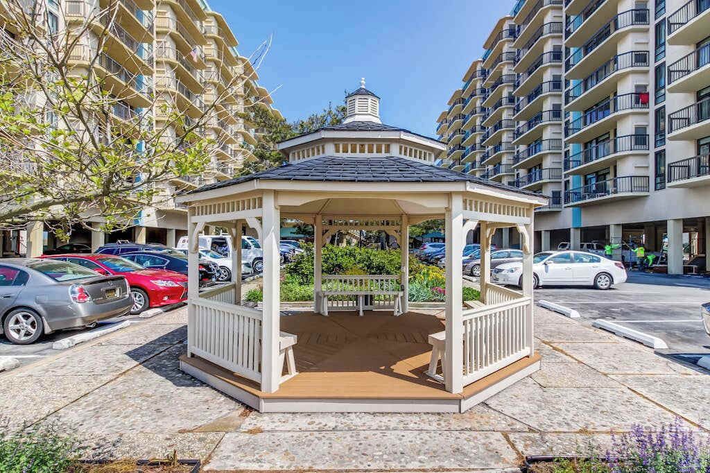 A gazebo with a wooden bench, surrounded by a paved area with parked cars and a multi-story building in the background.