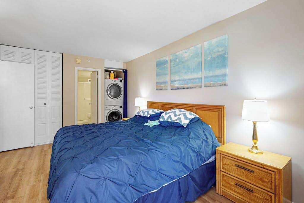 A bedroom with a bed, a nightstand, a lamp, a washing machine, and a closet.