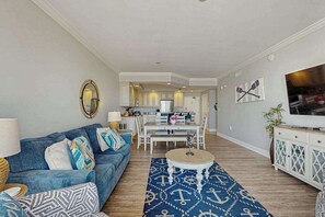 A living room with a sofa, coffee table, and a patterned rug. There is a dining area with a table and chairs, and a kitchen in the background. - Belmont Towers 410 (Ocean City)
