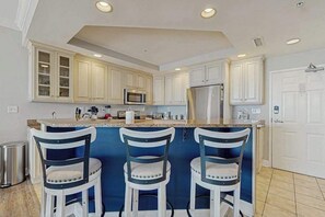 A kitchen with white cabinets, a central island with bar stools, a stainless steel refrigerator, and a microwave. - Belmont Towers 410 (Ocean City)