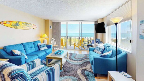 Un salón con un sofá azul, una mesa de centro y un televisor. Hay una ventana grande con vista al mar.