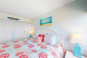 A bedroom with a bed, a nightstand, a lamp, a painting, and a surfboard decoration. - Gorgeous oceanfront condo with pool, central AC, WiFi, balcony water views (Ocean City)
