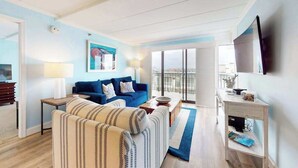 A modern living room with a sofa, a coffee table, a TV, and a balcony with a view. - Beachfront Condo w/Free WiFi, AC, Private Washer/Dryer, Shared Pool, Water Views (Ocean City)