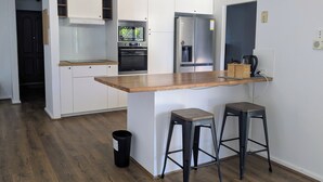 A modern kitchen with white cabinetry, a wooden countertop, a built-in oven, a refrigerator, and two stools. - Welcoming 3-Bed House with Large Backyard (Mandurah)