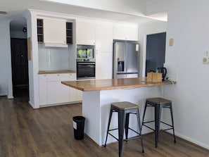 A modern kitchen with white cabinetry, a wooden countertop, a built-in oven, a refrigerator, and two stools. - Welcoming 3-Bed House with Large Backyard (Mandurah)
