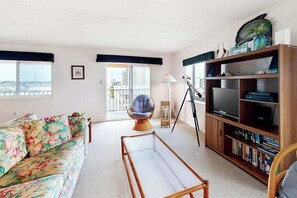 Ein Wohnzimmer mit einer Couch, einem Sessel, einem Bücherregal, einem Fernseher und einem Balkon mit Aussicht.