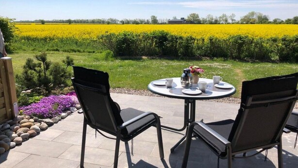 Un patio exterior con dos sillas y una mesa, un campo de flores amarillas al fondo y un cielo despejado.