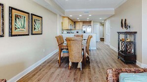 A dining area with a central table and chairs, a kitchen in the background, and framed artwork on the walls. - Condo in an oceanfront building with side ocean view, pool, gym & garage parking (Ocean City)