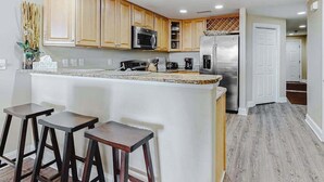 A kitchen with wooden cabinets, a stainless steel refrigerator, a microwave, and a coffee maker. There are three wooden stools. - Condo in an oceanfront building with side ocean view, pool, gym & garage parking (Ocean City)