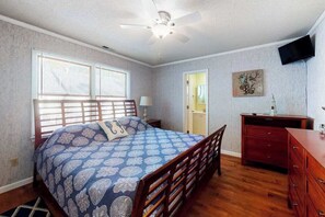 Une chambre à coucher comprenant un lit, une commode en bois, un ventilateur de plafond et un téléviseur fixé au mur.