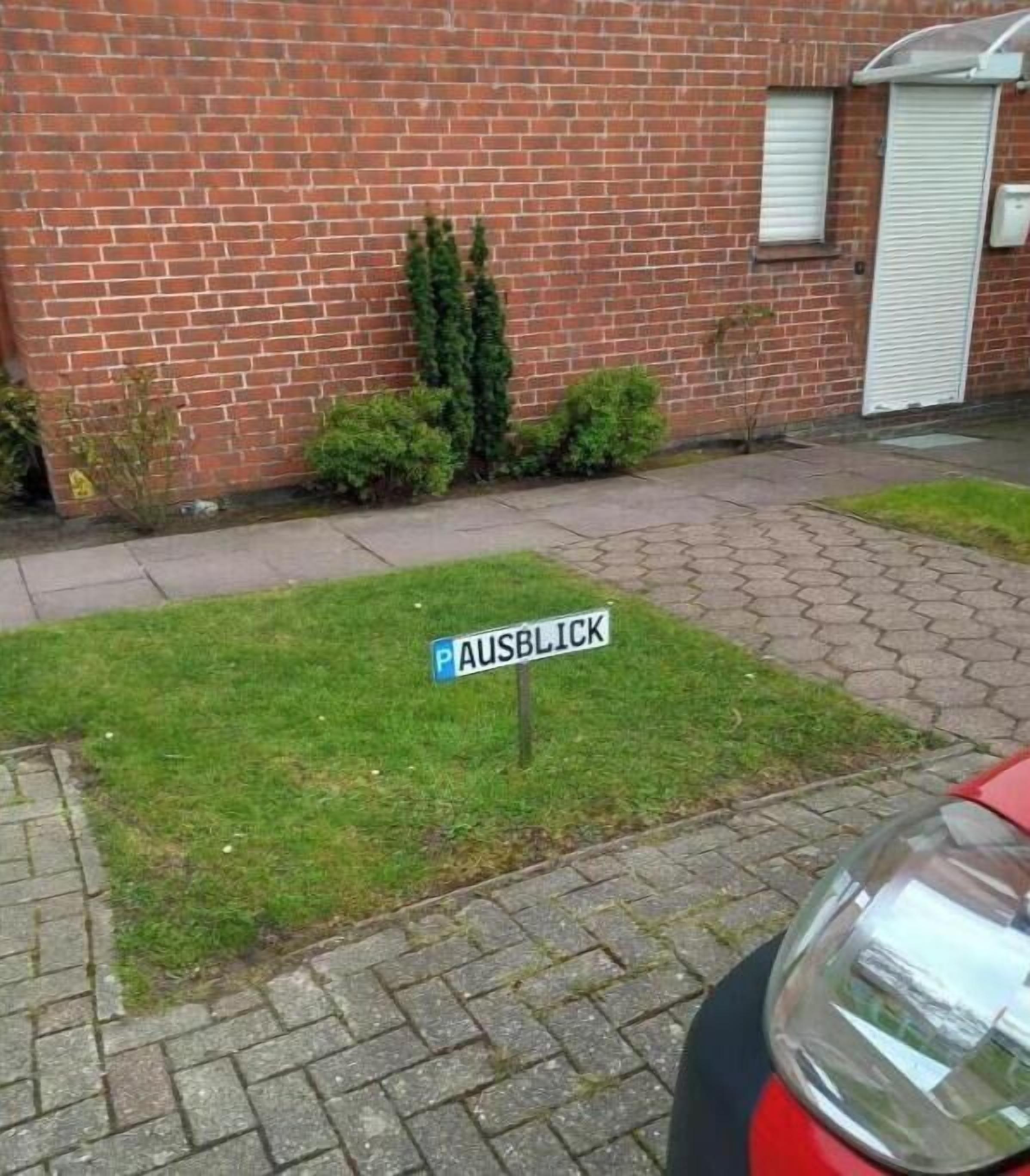 A street sign labeled 'AUSBLICK' in front of a brick building with a small lawn and a red car parked nearby.